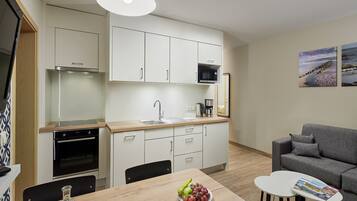 Et moderne køkken med hvide skabe, en mikrobølgeovn og en vask. Der er et spisebord med en skål frugt og en glasflaske. I opholdsstuen er der en grå sofa og et lille hvidt bord med et magasin.
