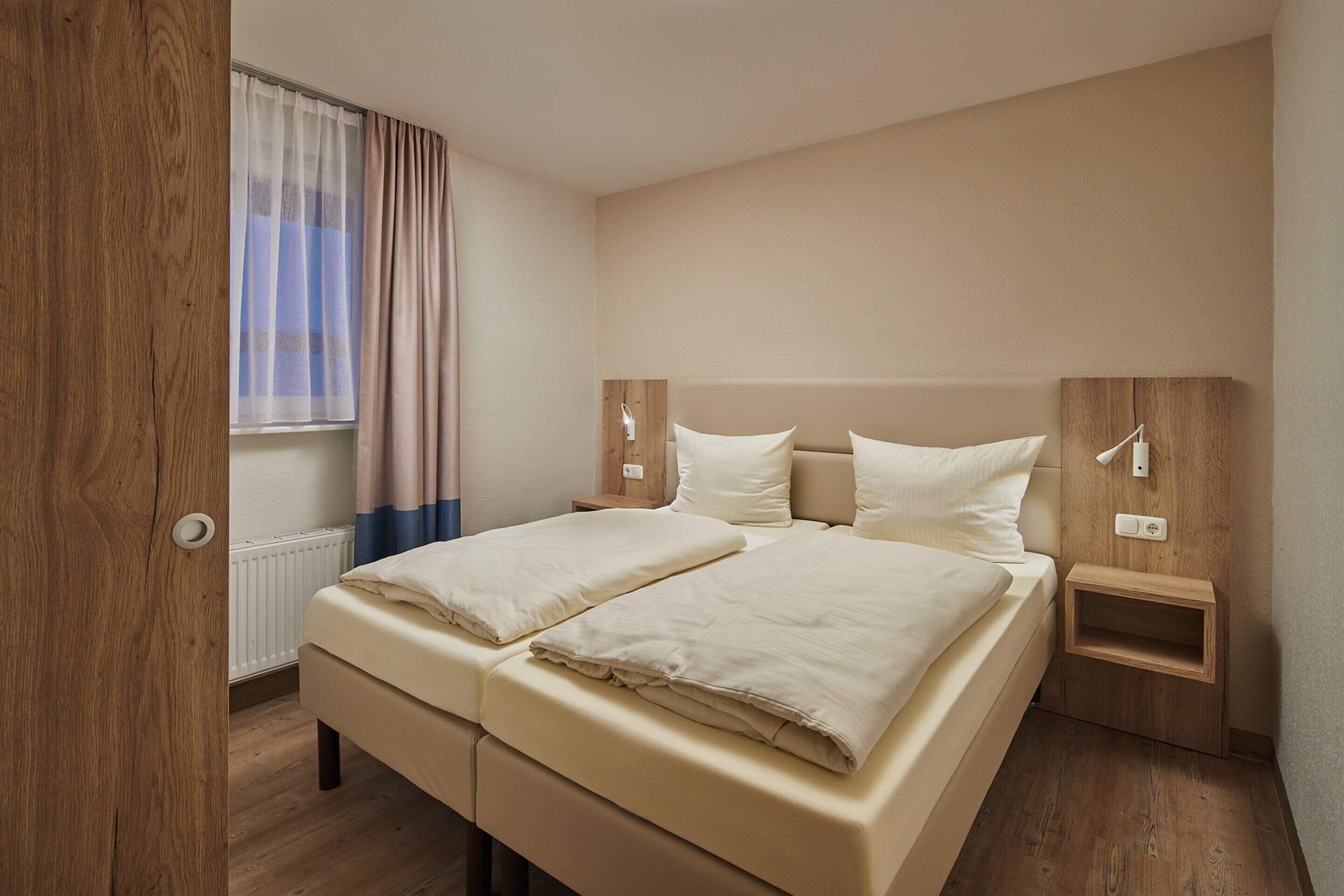 Ein Schlafzimmer mit Doppelbett, Holz-Kopfteil, Nachttischen und einem Fenster mit Vorhängen.