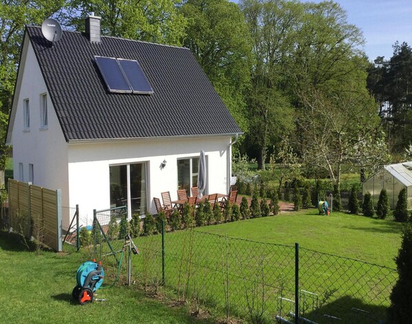 Une maison avec un toit en pente, des panneaux solaires, une cour clôturée et une serre.