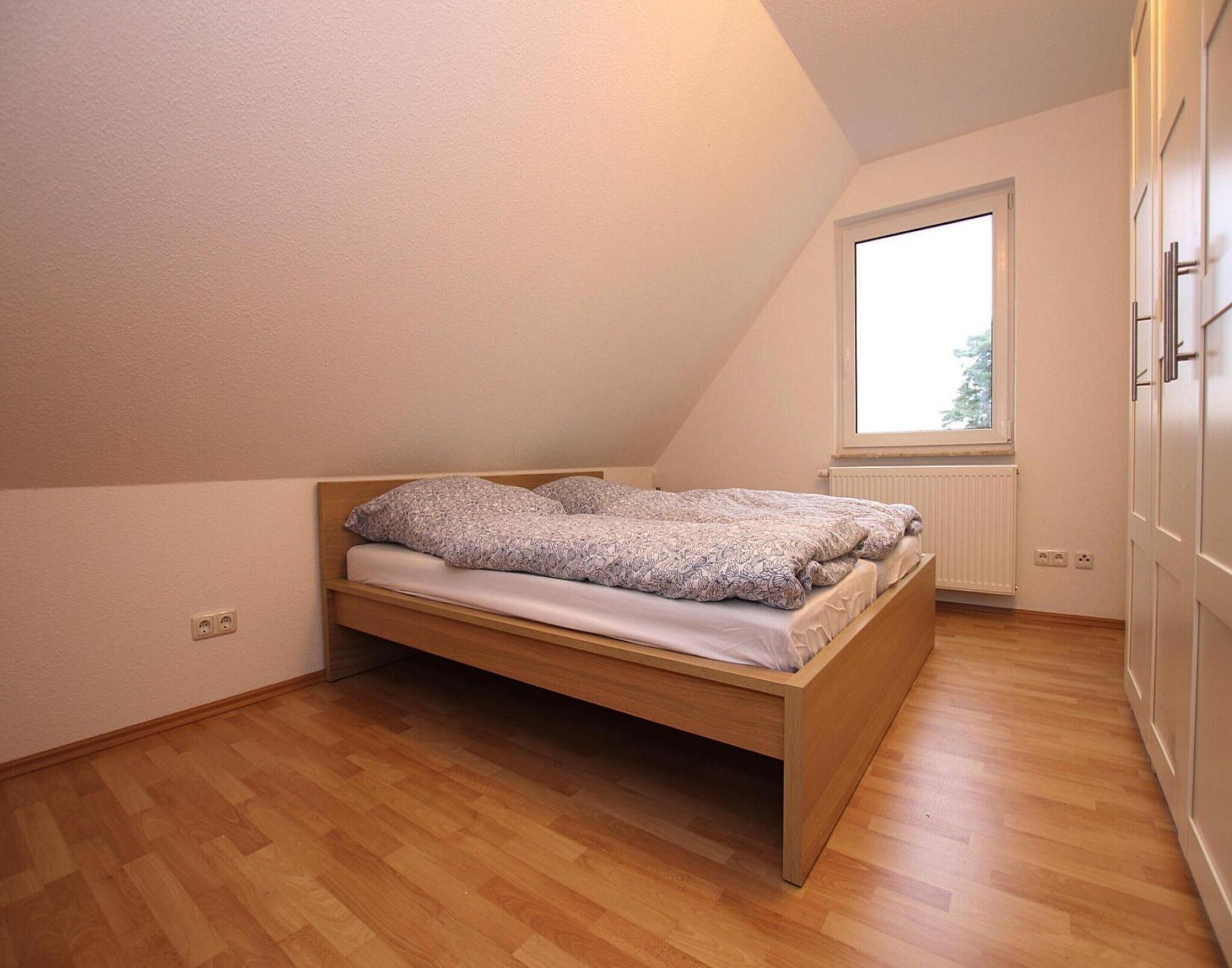 Een kleine kamer met een enkel bed en schuine wand, houten vloer, een raam en een radiator.