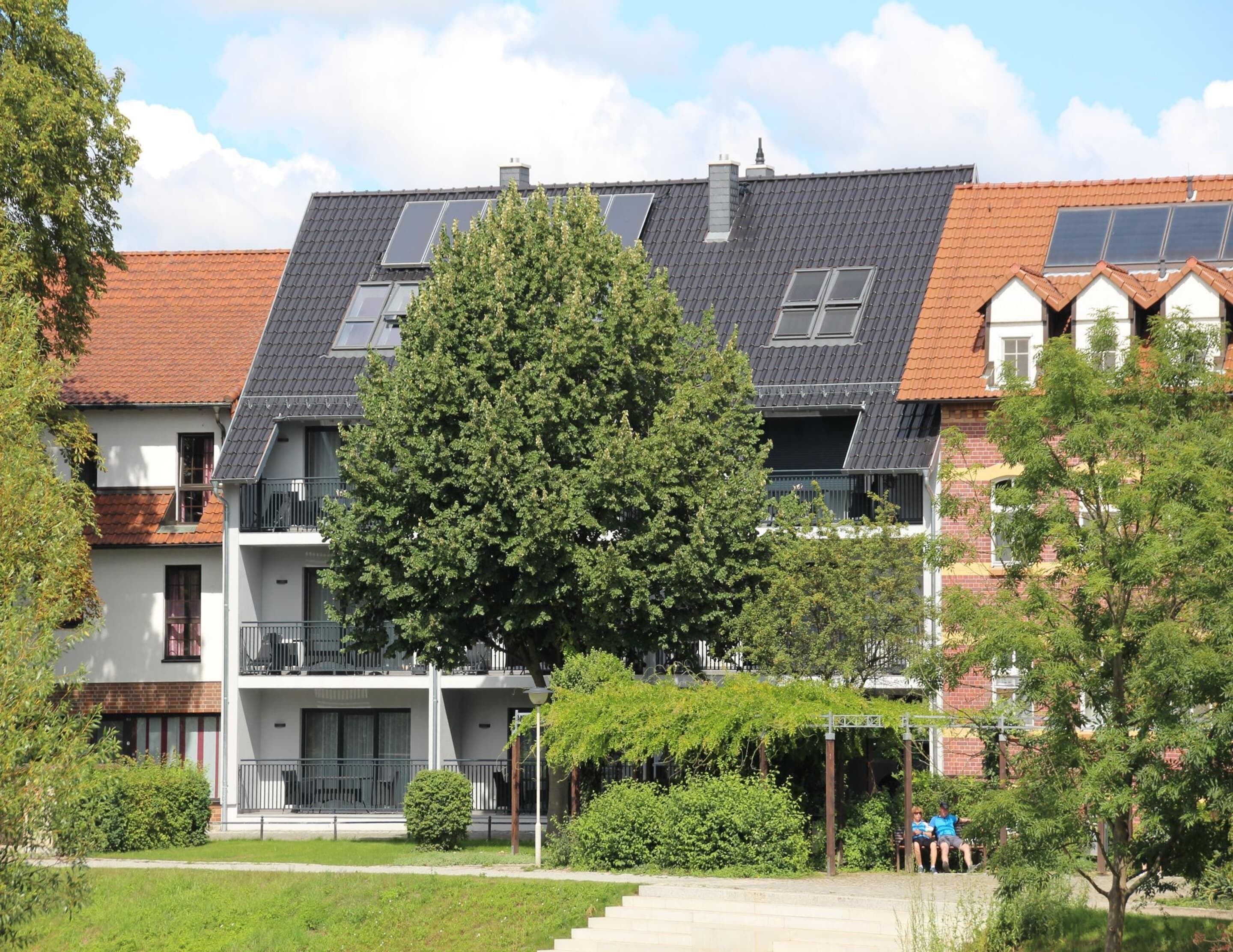 Un edificio residencial con techo inclinado, paneles solares y un árbol grande en la fachada.