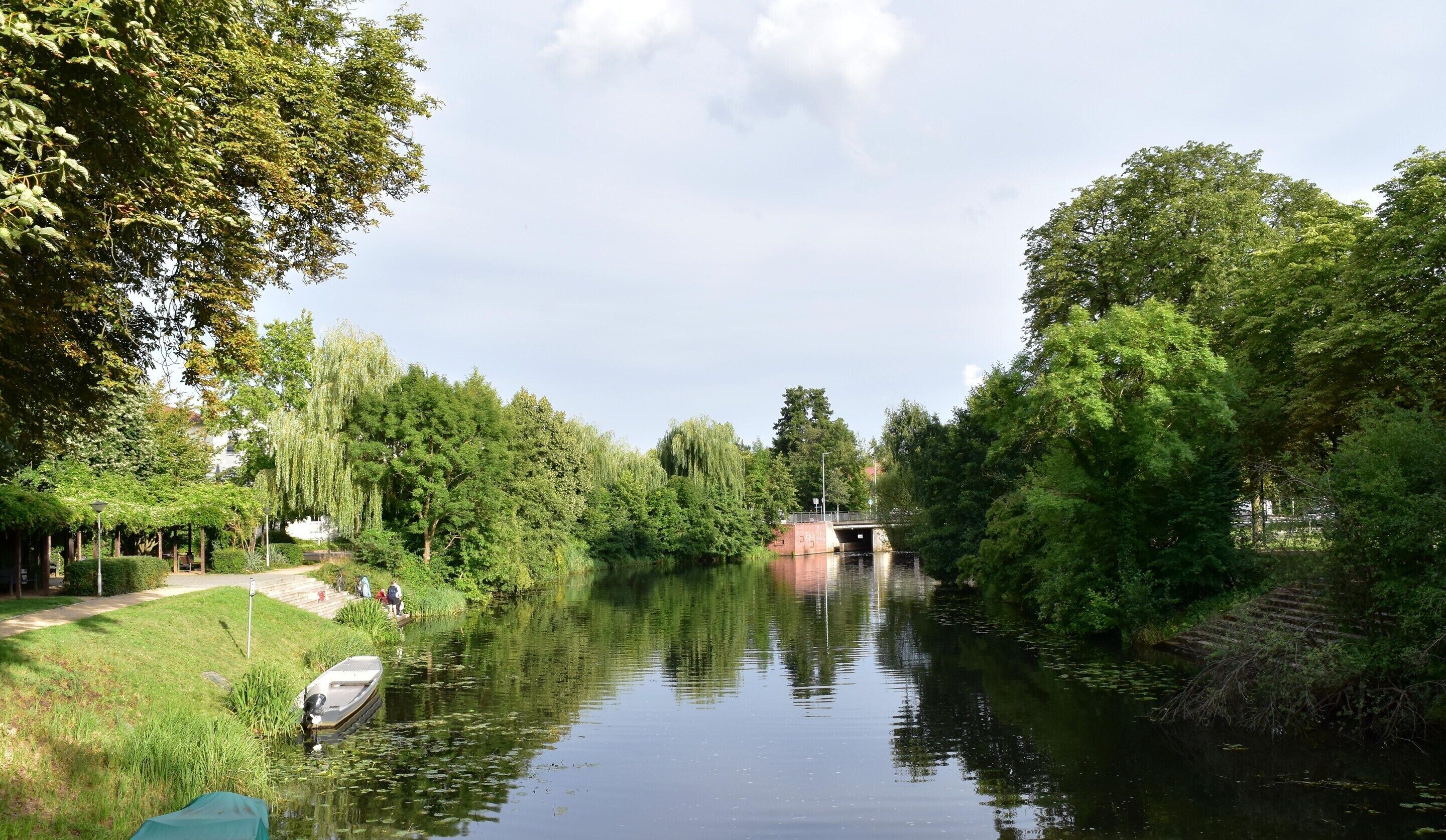 Une rivière paisible, traversée par un pont, bordée d’une végétation luxuriante, et un bateau amarré sur la rive gauche.