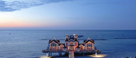 Un hotel frente al mar con un muelle largo que se adentra en el océano, que cuenta con varias habitaciones y un edificio central con techo a dos aguas.