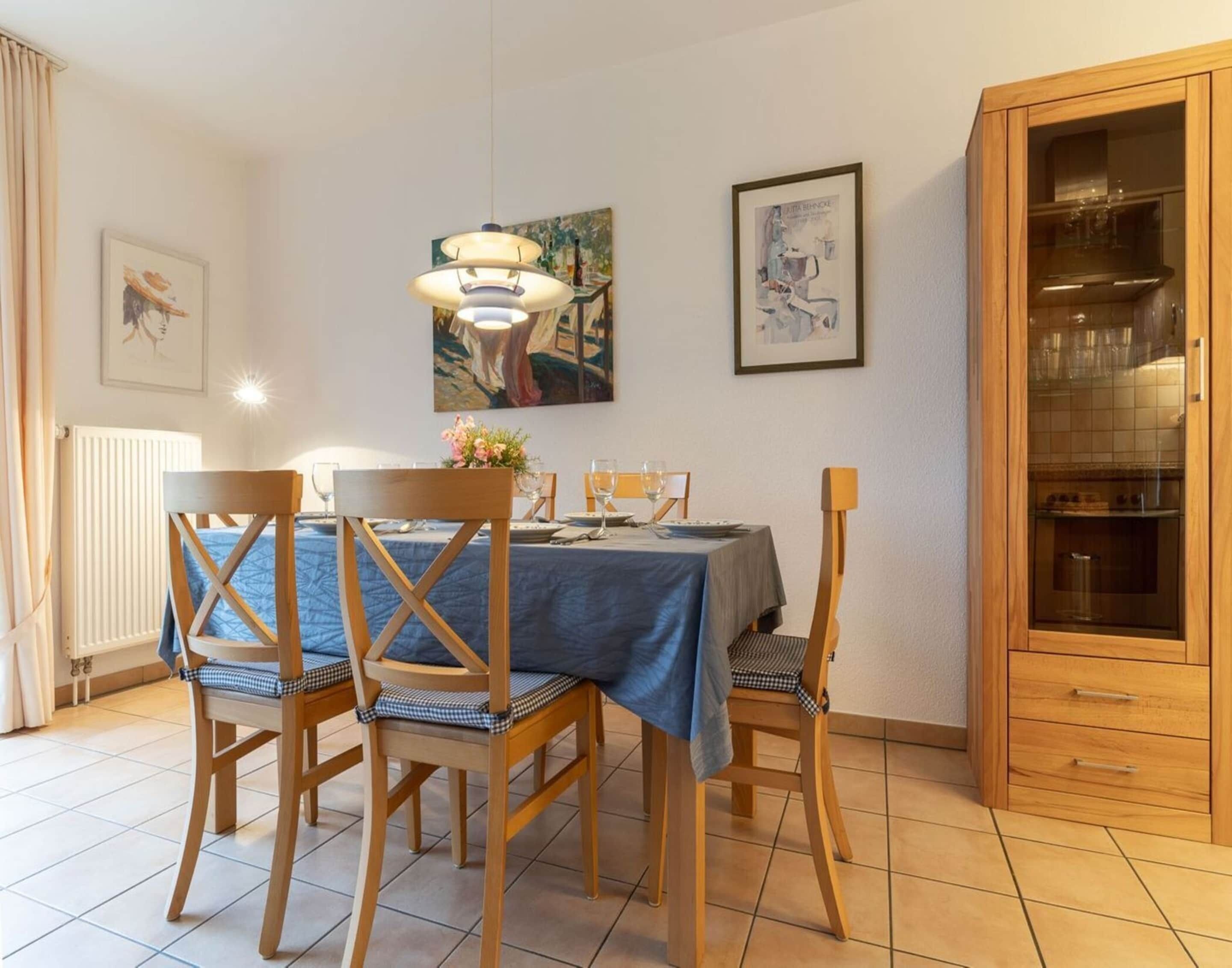 A dining area with a table set for four, wooden chairs with checkered cushions, a blue tablecloth, and a hanging light fixture.
