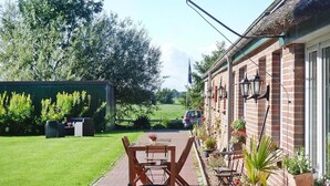 A patio area with a wooden table and chairs, potted plants, and a brick building with windows and outdoor lighting. - Ferienhof Landhaus Markus, Suderschwei (Stadland)