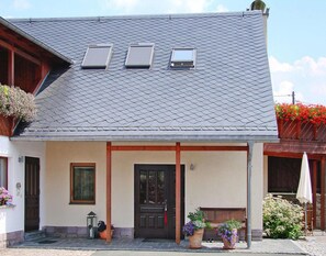 Uma casa de dois andares com telhado de telhas cinzas, porta da frente e varanda com um banco.