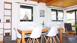 Un espace repas avec une table en bois et des chaises blanches, de grandes fenêtres avec des rideaux, un plafond en bois et un fauteuil en osier.