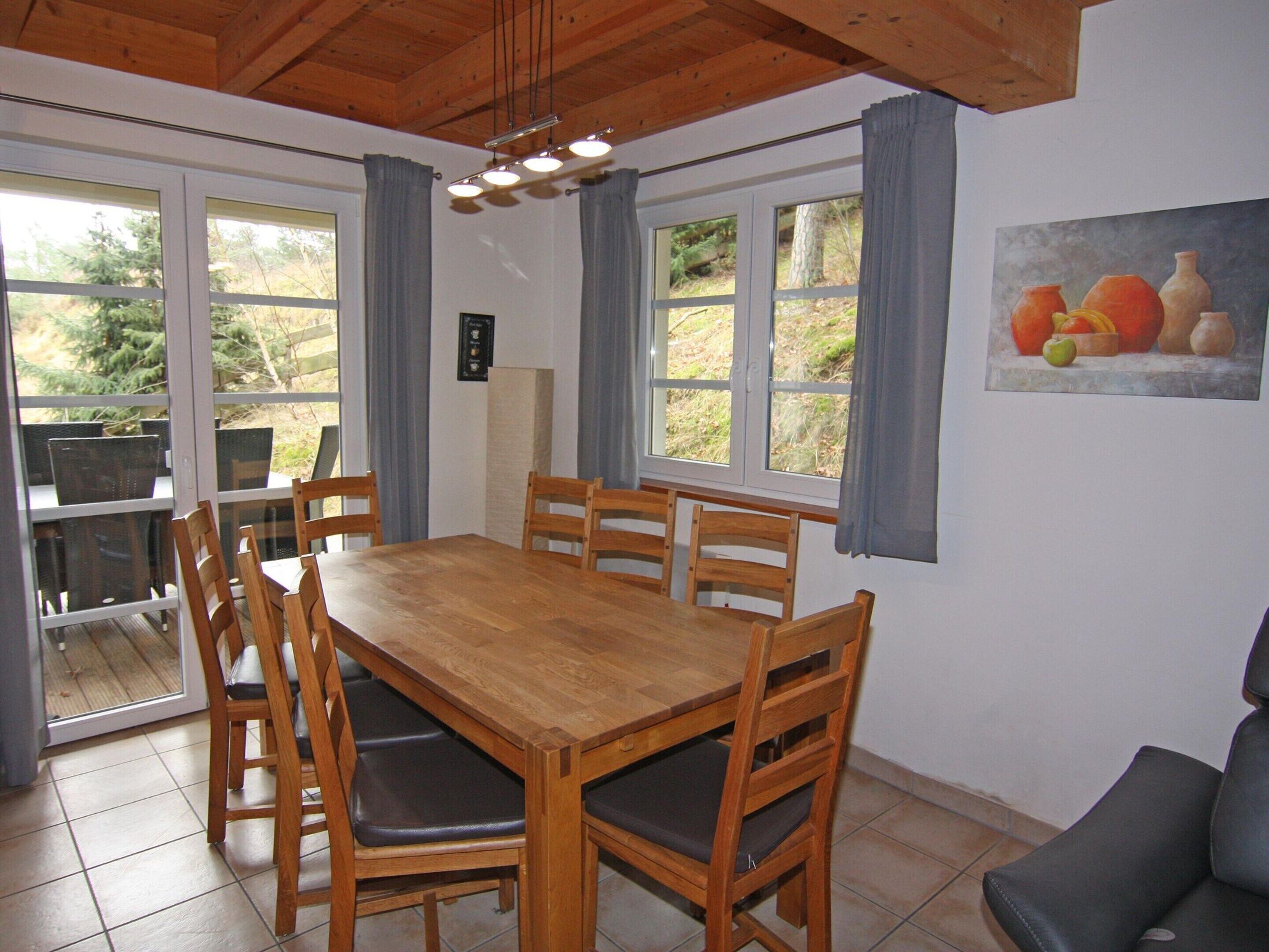 Un comedor con una mesa y sillas de madera, ventanas amplias con cortinas y un cuadro de frutas y verduras en la pared.