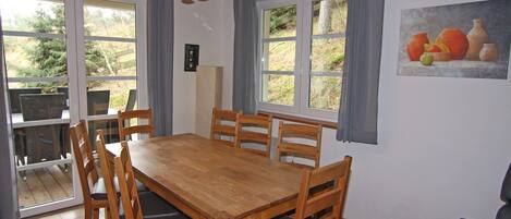 Un comedor con una mesa y sillas de madera, ventanas amplias con cortinas y un cuadro de frutas y verduras en la pared.