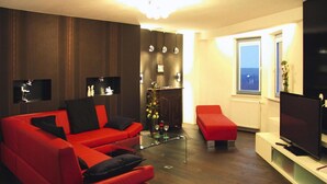 En moderne stue med en rød sofa, en lille rød skammel, et glasbord og et fladskærms-tv på et hvidt skab.