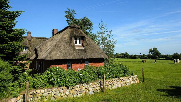 Ein traditionelles Haus mit Strohdach, einer Steinmauer und einem grünen Feld im Hintergrund.