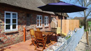 Ein Haus aus Ziegelsteinen mit einem Strohdach, einer Terrasse mit Tisch und Stühlen sowie einem Holzzaun.