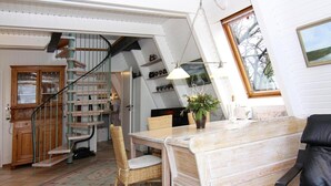 A cozy room with a wooden dining table, wicker chairs, a black leather sofa, and a spiral staircase leading to a loft area with shelves. - Ferienhaus in Damp mit Terrasse (Damp)