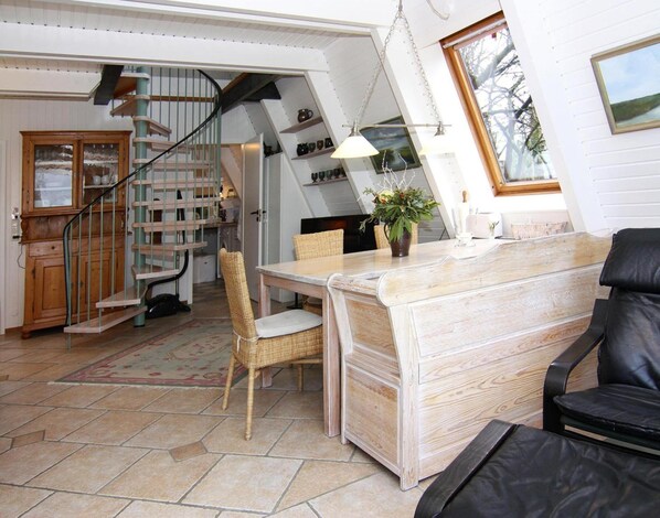A cozy room with a wooden dining table, wicker chairs, a black leather sofa, and a spiral staircase leading to a loft area with shelves.