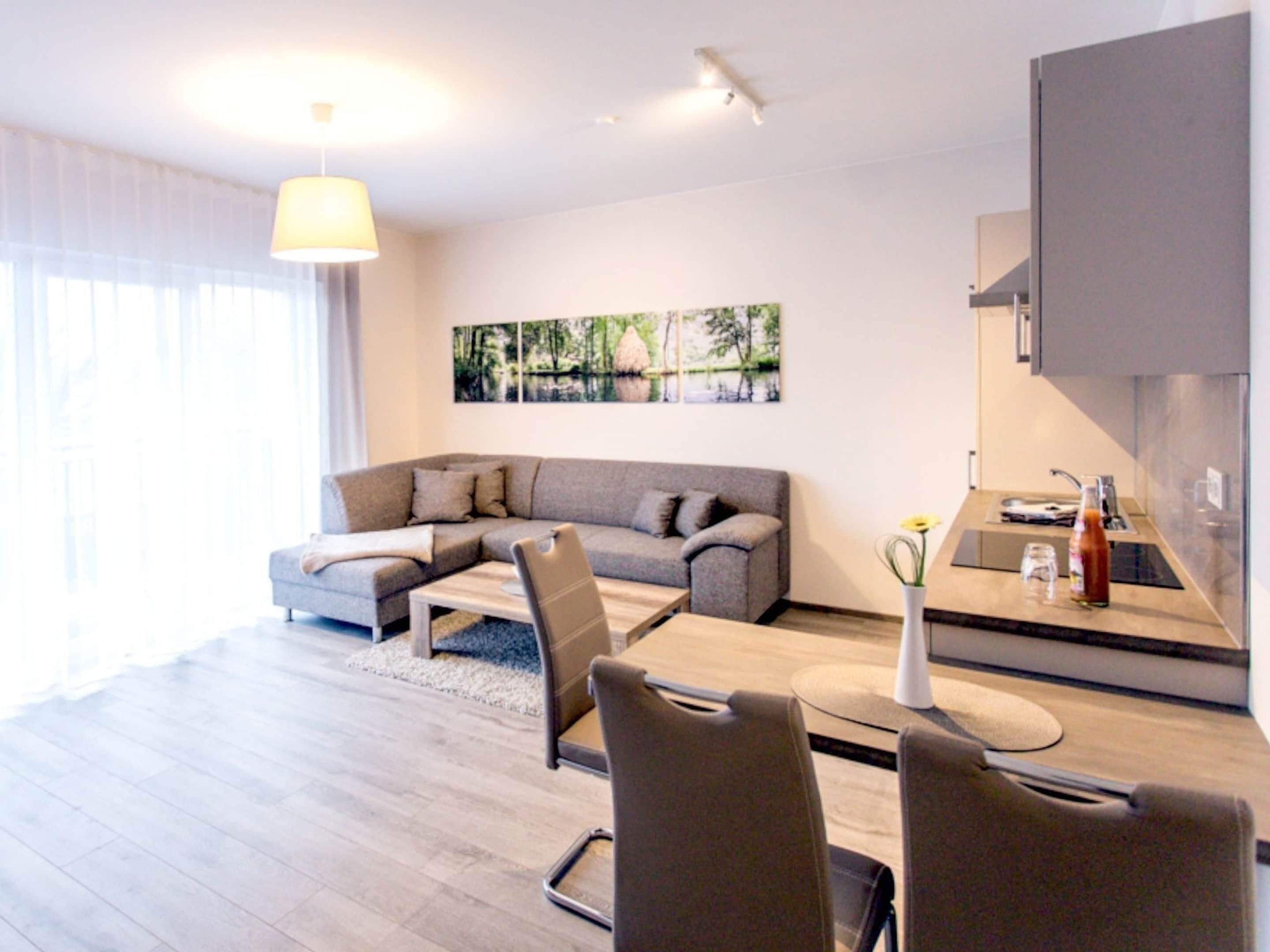 Un salon moderne avec un canapé d’angle gris, une table basse en bois et un coin repas comprenant une petite cuisine.