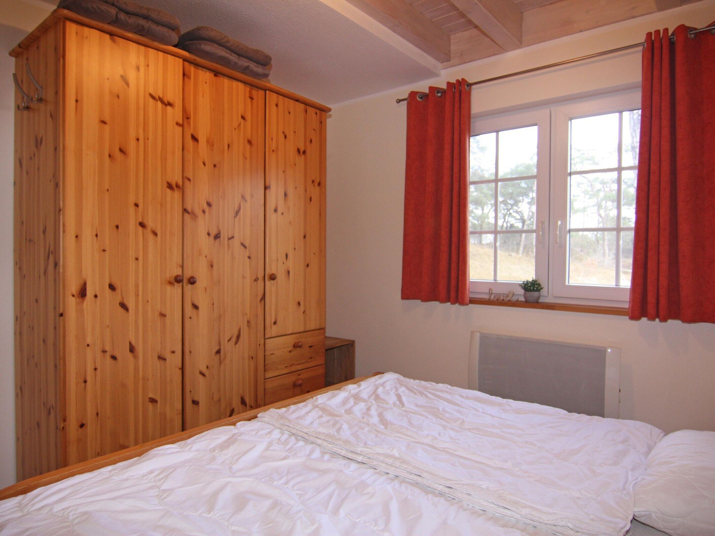 A bedroom with a wooden wardrobe, a bed with white bedding, a window with red curtains, and a radiator.
