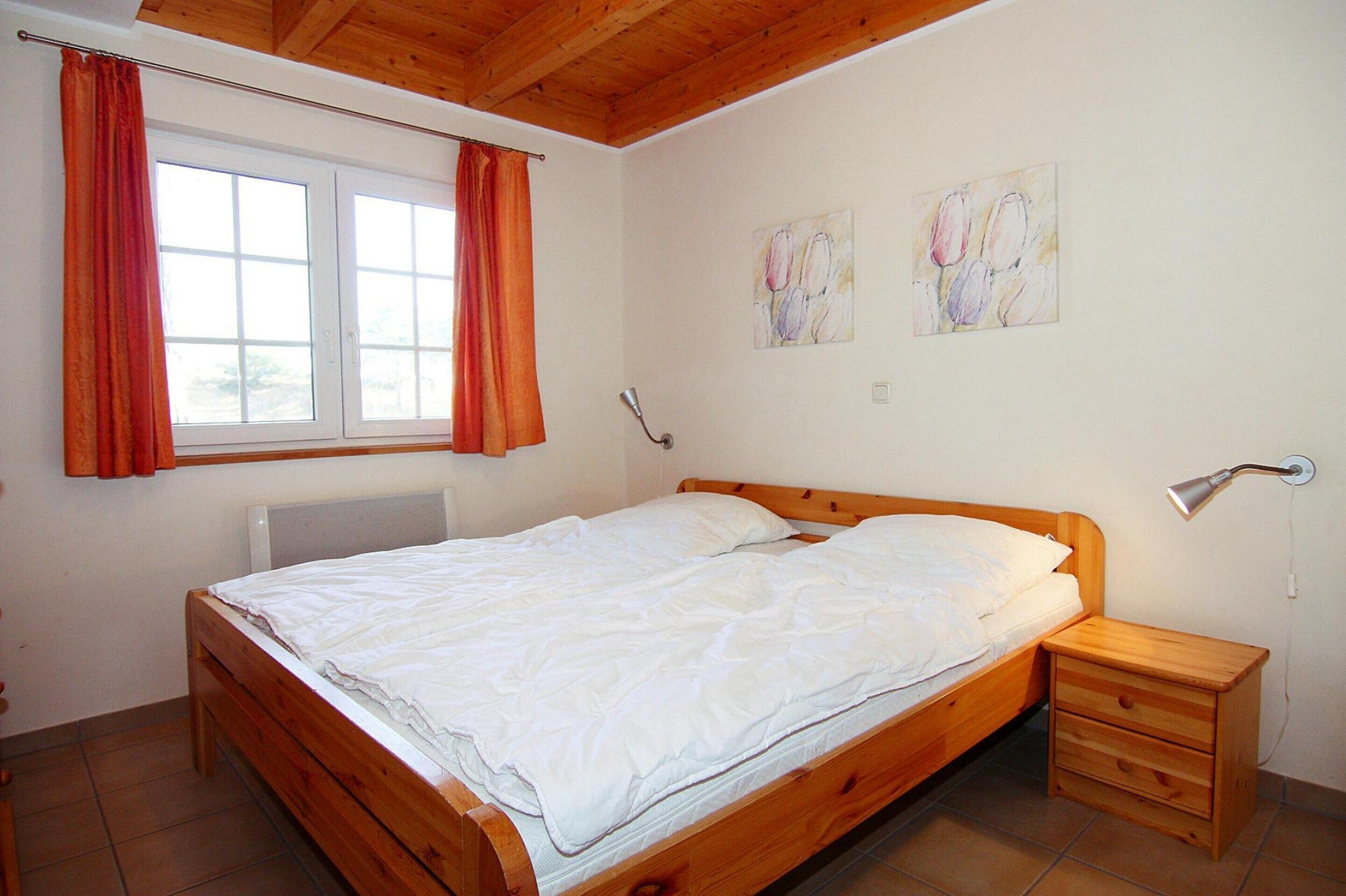 Ein Schlafzimmer mit einem hölzernen Bett, einem Nachttisch, einem Fenster mit Vorhängen und zwei an der Wand befestigten Bildern.