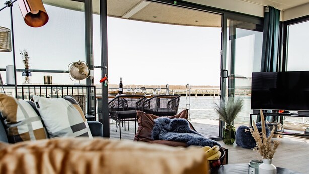 Un salón moderno con un sofá, una mesa de centro y un televisor. Se ve el agua a través de amplias ventanas.
