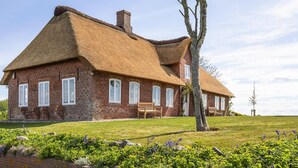 Une maison en briques avec un toit de chaume, un banc et un arbre au premier plan.