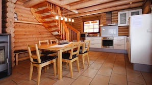 Interior de uma cabana de madeira com uma mesa de jantar, cadeiras, cozinha, lareira e escada de madeira.