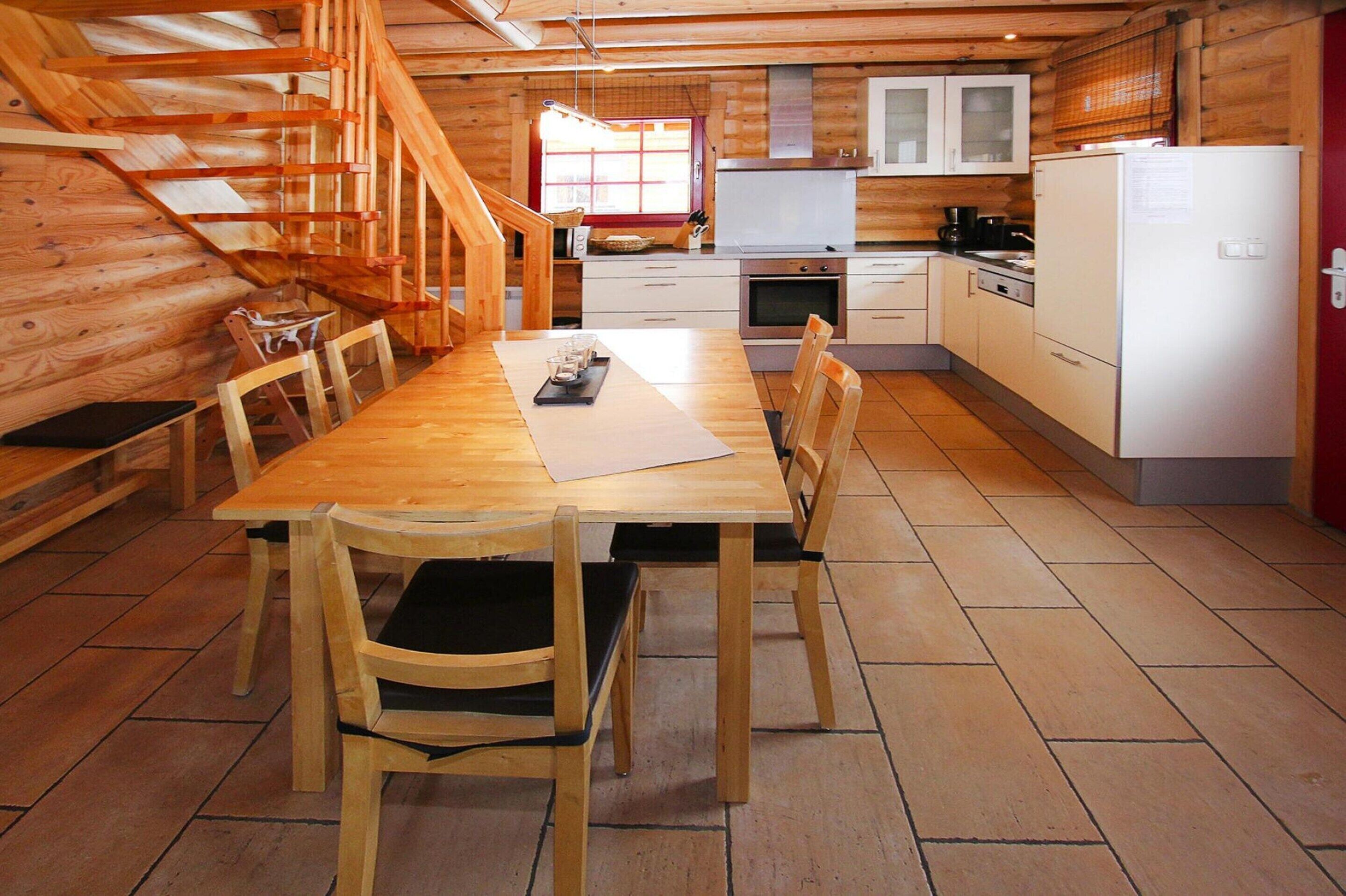 Interior de una cabaña de madera con una mesa de comedor, sillas, una cocina con armarios blancos y una escalera de madera.