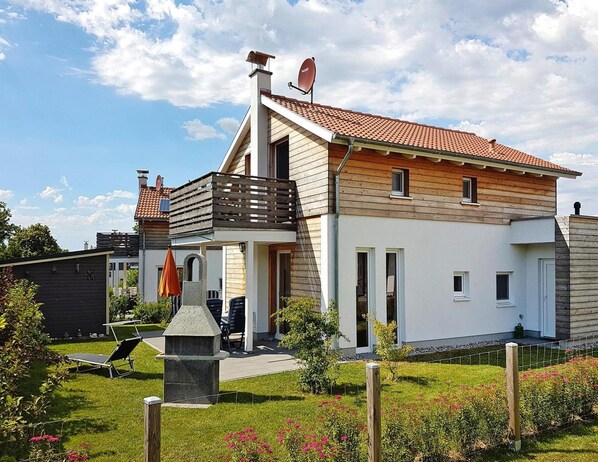Una casa a due piani con tetto spiovente, un balcone e un giardino fiorito con zona barbecue.