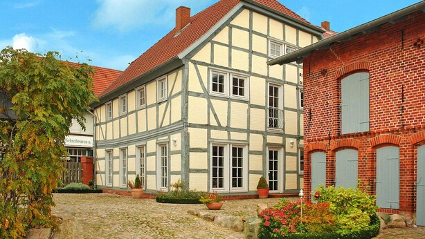 A traditional half-timbered house with a cobblestone courtyard, a brick building with a red roof, and a sign indicating a business. - Ferienhaus Rosenhof, Lenzen (Lenzen)