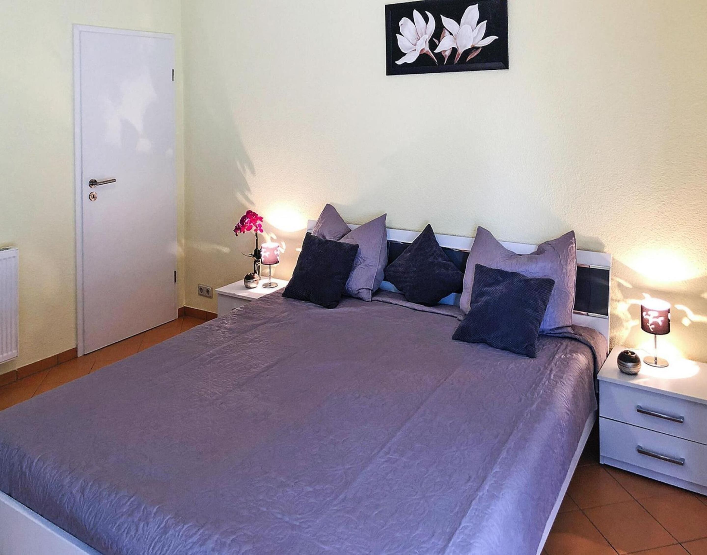 A bedroom with a large bed, a nightstand with a lamp, a door, and a wall-mounted picture.