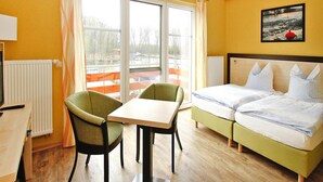 A hotel room with a bed, a desk, and a chair. There is a window with curtains, a radiator, and a picture on the wall. - Ferienwohnung Fewo Elde in Plau am See (Plau am See)
