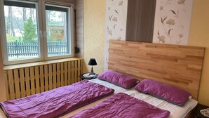Un dormitorio con cabecera de madera, dos camas con ropa de cama púrpura, una mesita de noche con lámpara y una ventana con vistas a la vegetación.