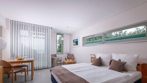 A modern hotel room with a large bed, a desk with a chair, a small table with a lamp, and a window with blinds. - Apartments, Gohren (Göhren)