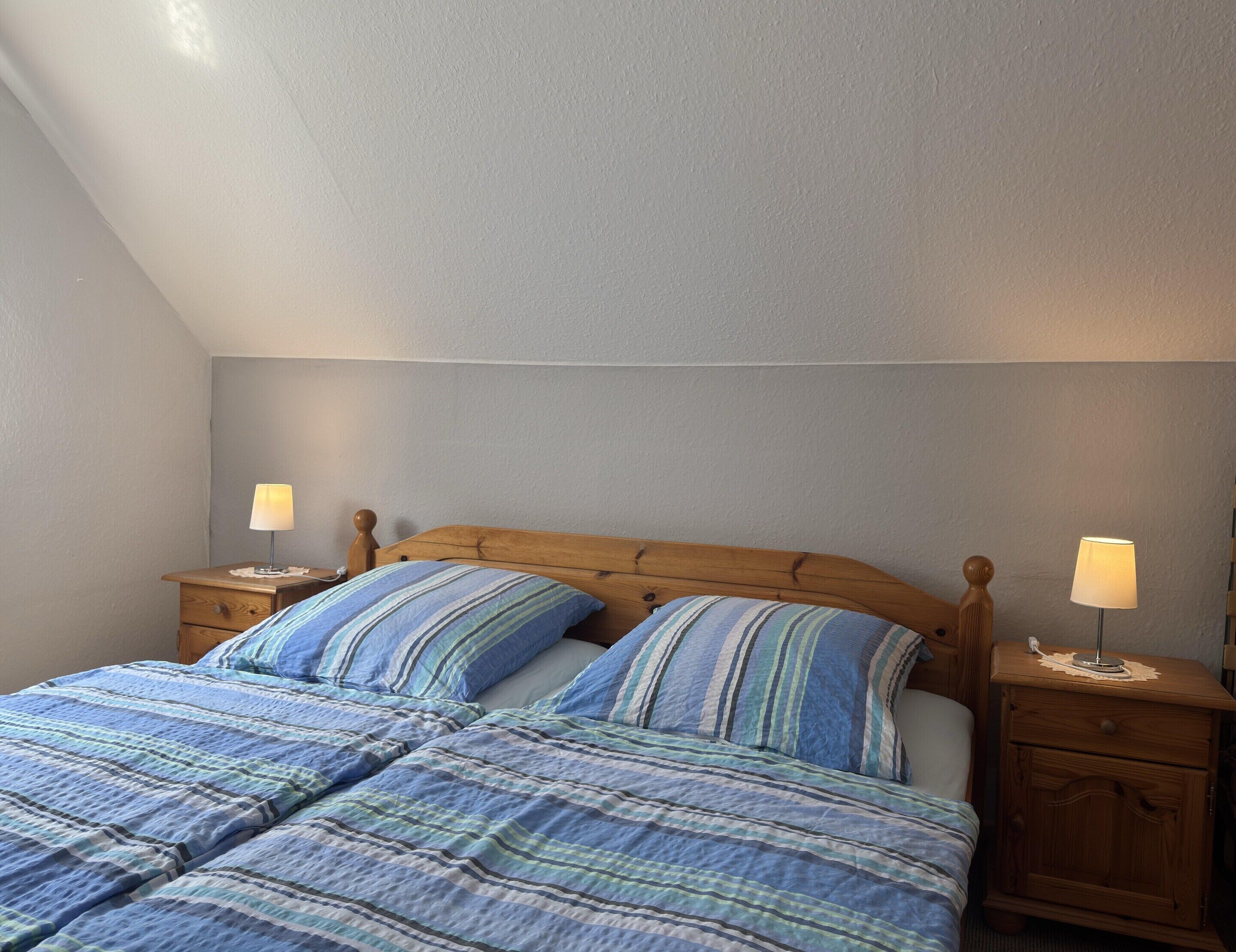 A bedroom with a sloped ceiling, a wooden bed frame, two bedside tables with lamps, and a striped bedspread.