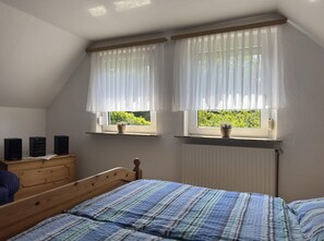A bedroom with a sloped ceiling, two windows with sheer curtains, a wooden dresser, a bed with a striped bedspread, and a radiator. - Ferienwohnung in Moorhusen (Sudbrookmerland)