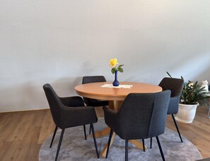 A round wooden table with a blue vase and a yellow flower, surrounded by four dark chairs with wooden legs. - Ferienwohnung in Moorhusen (Sudbrookmerland)