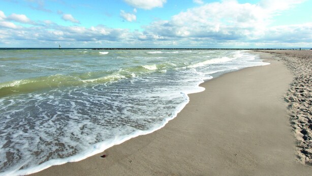 Uma praia com ondas suaves e céu claro, e um píer ao longe.