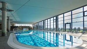 Uma grande piscina interior com água azul cristalina, rodeada por amplas janelas com vista para a cidade.