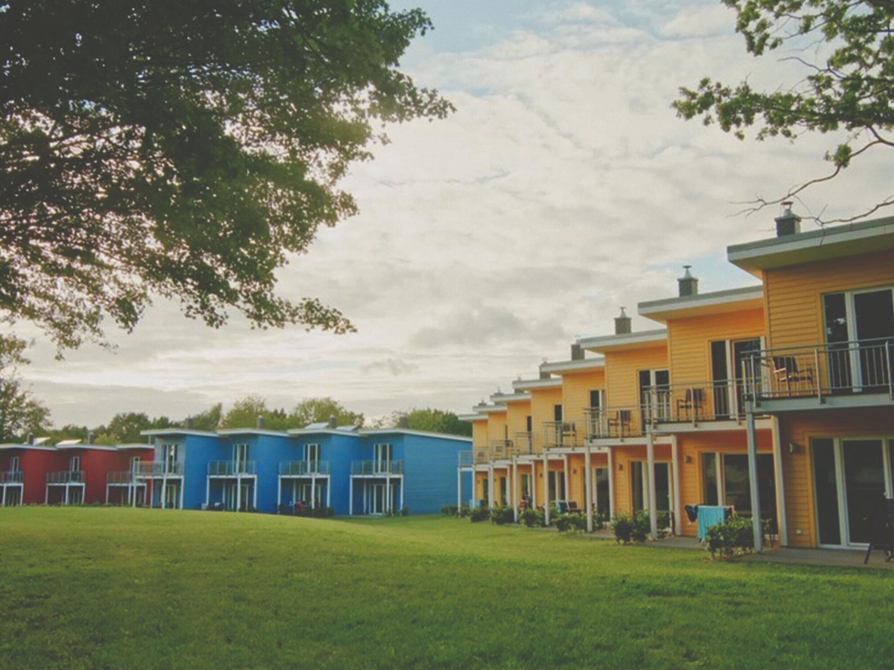 A row of colorful single-story buildings with balconies, set against a backdrop of trees and a cloudy sky.