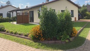 Einbungalow mit überdachter Terrasse, umzäunter Garten und einem Blumenbeet mit gelben Blüten.