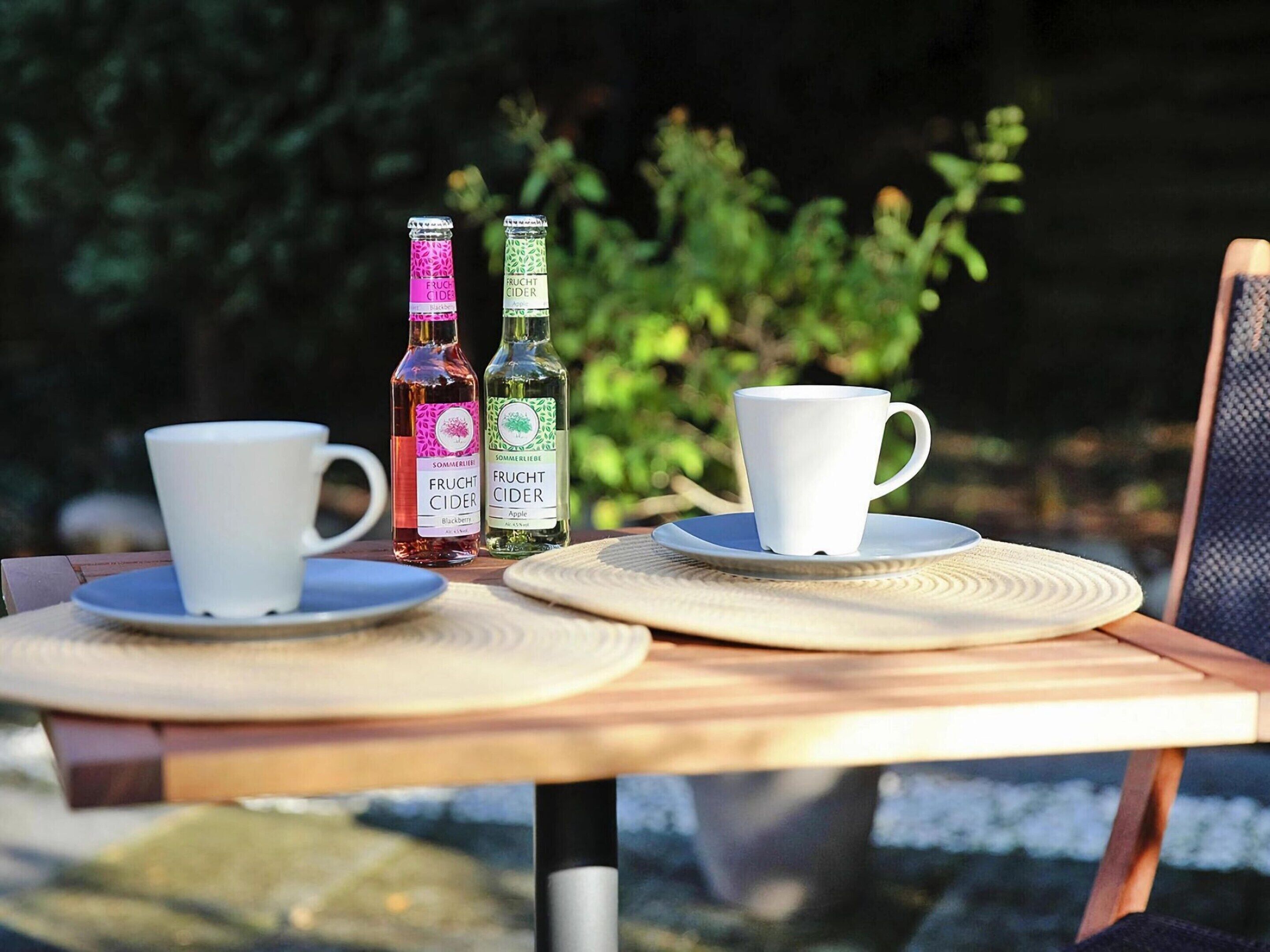 A table with two cups and two bottles of fruit cider, set outdoors with greenery in the background.