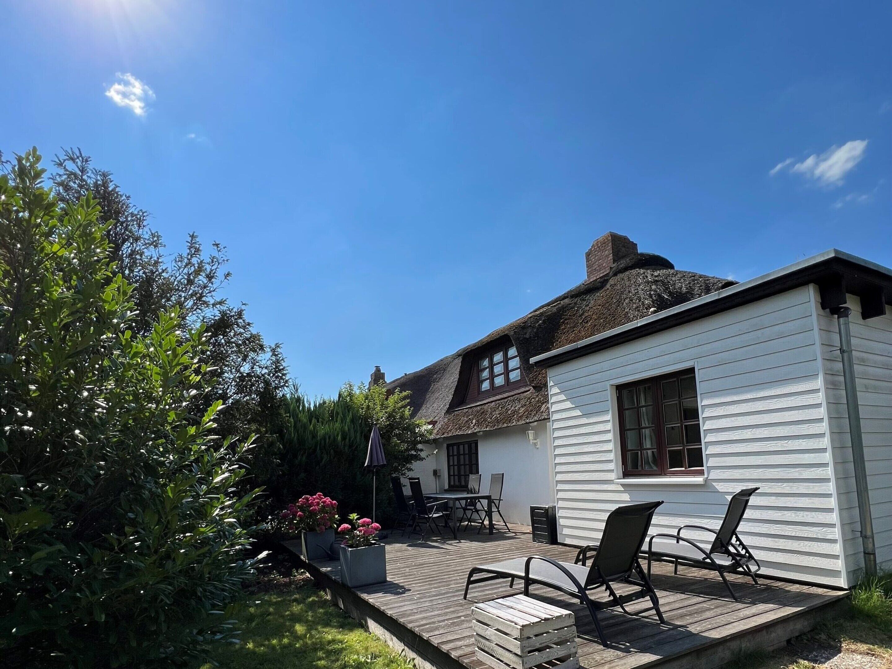 A white house with a thatched roof, a wooden deck with two chairs, and a garden with green shrubs and pink flowers.