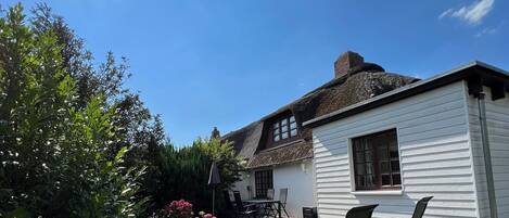 A white house with a thatched roof, a wooden deck with two chairs, and a garden with green shrubs and pink flowers.