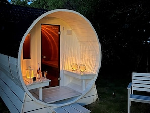 Une petite structure circulaire en bois, accueillante, avec une porte, une petite table sur laquelle sont posés des verres et des bouteilles, et un banc à l’extérieur.