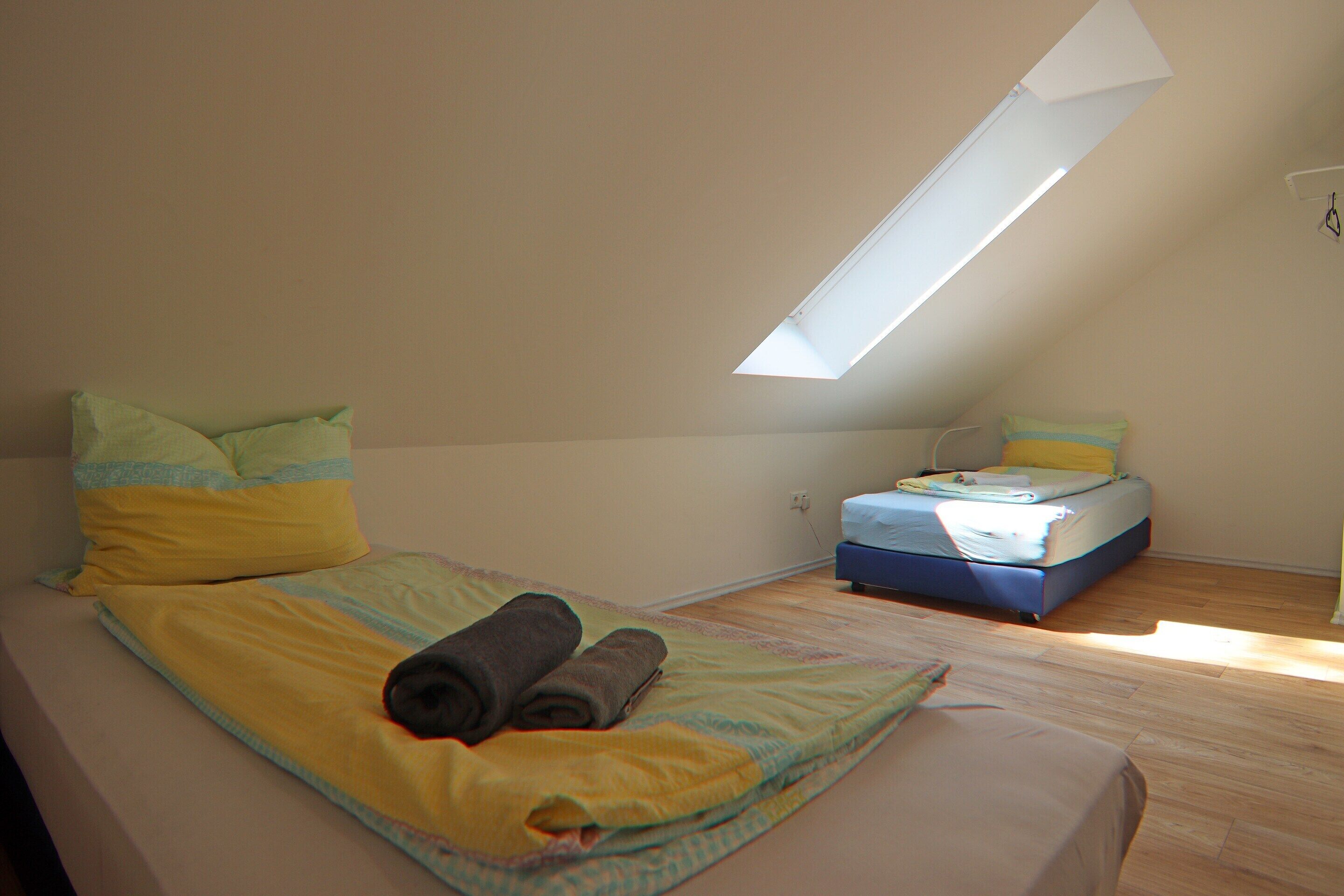 Una camera da letto con soffitto spiovente, una finestra a lucernario, un letto con un asciugamano arrotolato e un materasso a terra.