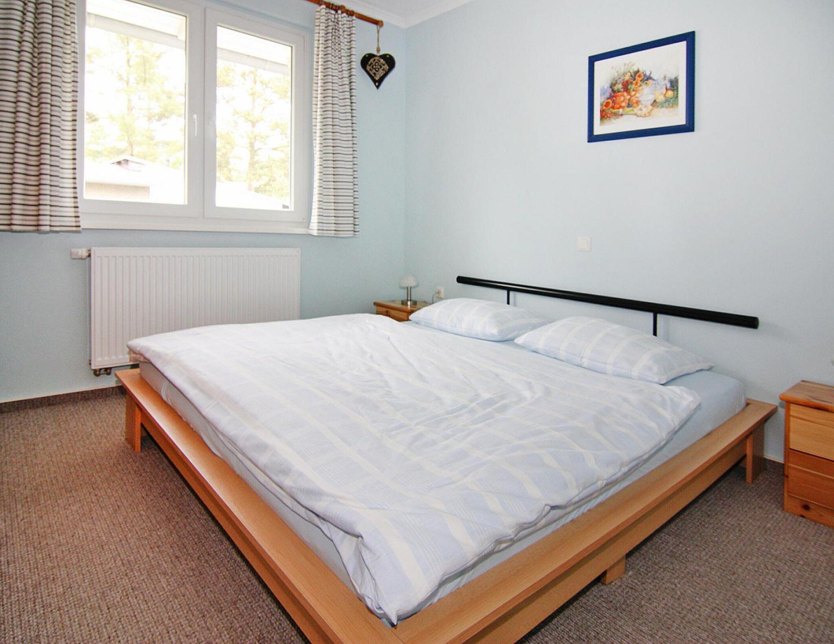 Habitación con cama individual de estructura de madera, colchón blanco y cabecero negro. Tiene una ventana con persianas, un radiador y una mesita de noche con lámpara.