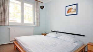 A single-bed room with a wooden frame, a white mattress, and a black headboard. There is a window with blinds, a radiator, and a small bedside table with a lamp. - Appartementhaus, Zinnowitz (Zinnowitz)