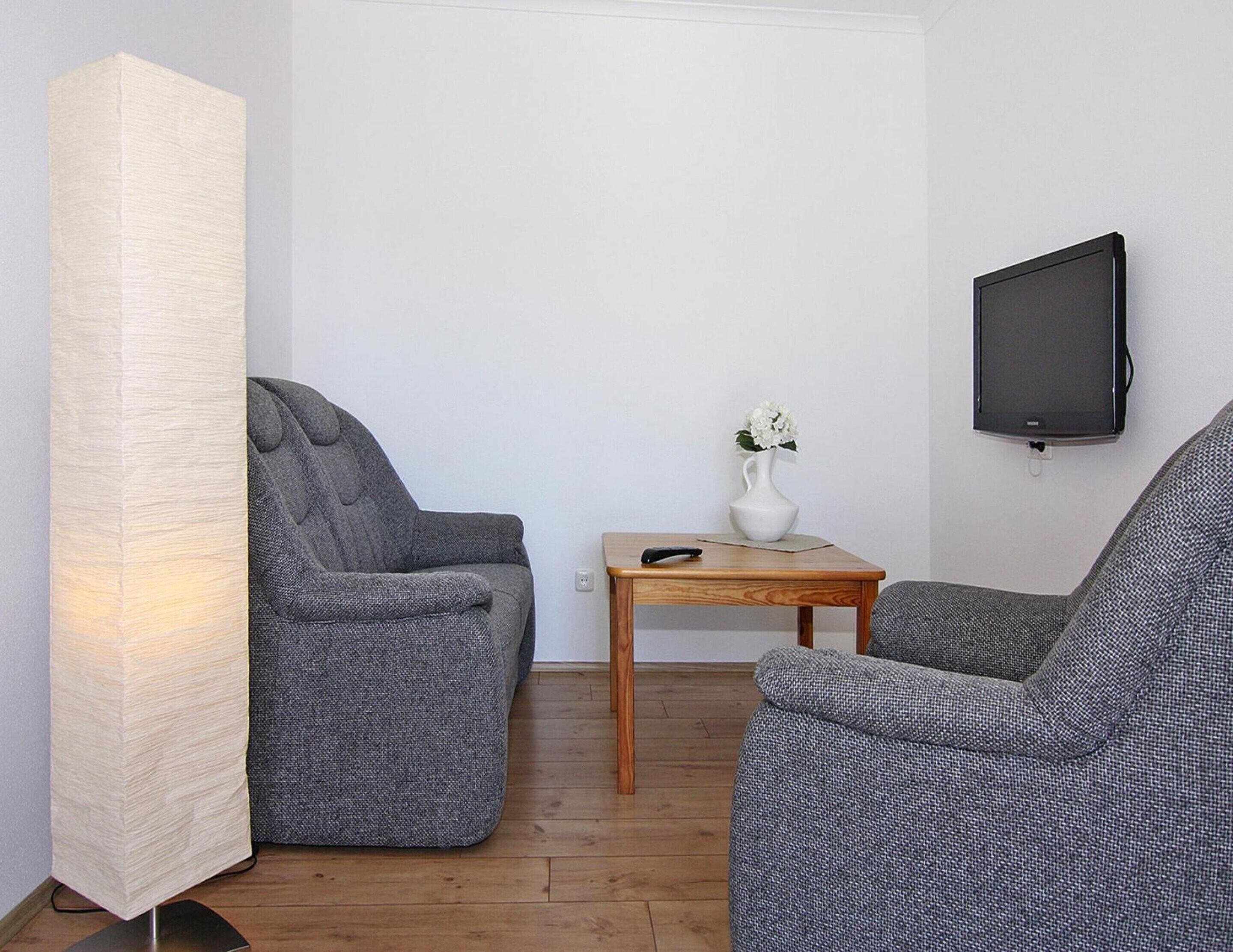 Un salon avec deux fauteuils gris, une petite table en bois sur laquelle est posé un vase de fleurs, un lampadaire et un téléviseur fixé au mur.