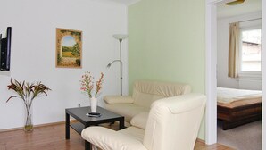 A room with a beige armchair, a small table with a vase of flowers, a painting on the wall, and a bed in the background. - Appartementhaus, Zinnowitz (Zinnowitz)