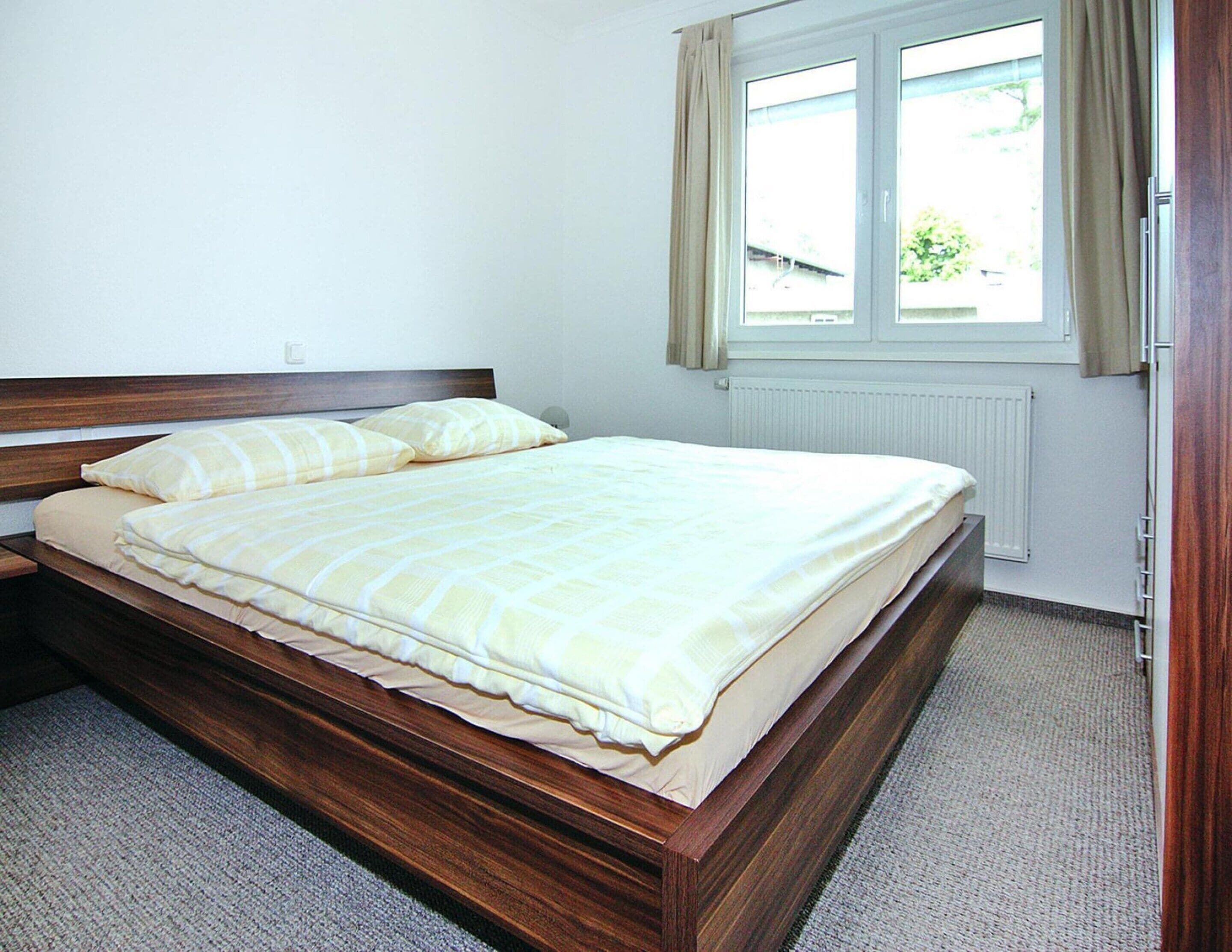 Ein ordentlich bezogenes Bett mit massivem Holzkopfstück in einem Zimmer mit Fenster und Heizkörper.