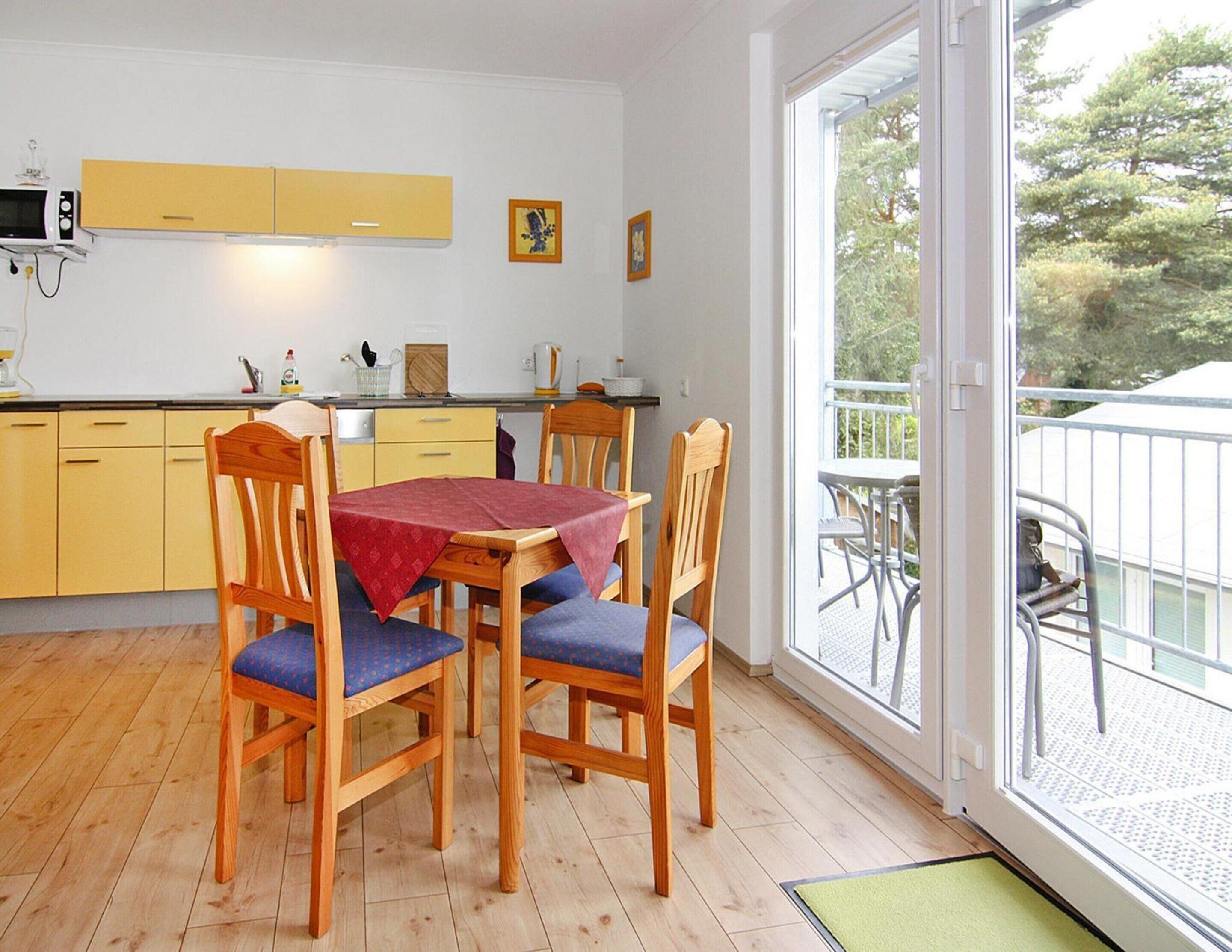 Et spiseområde med et træbord og stole, et gult køkken i baggrunden og en balkon med havemøbler.
