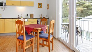 A dining area with a wooden table and chairs, a yellow kitchen in the background, and a balcony with outdoor furniture. - Appartementhaus, Zinnowitz (Zinnowitz)